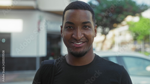 Canvastavla Young black man smiling with visible teeth and backpack strap, facing camera on street near parked car and storefront; approachability confidence