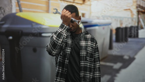 Φωτογραφία Man holds wrench to forehead on a street beside large metal dumpsters and trash bins in an urban alley; weariness repair struggle