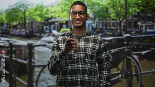 Foto Man holding a corrugated paper coffee cup in hand and looking down at it while standing on a city street beside a canal and parked bicycles; contemplation
