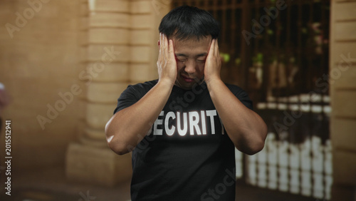 Foto Man security guard wearing black tshirt covers face with hands at historic building entrance; anxiety