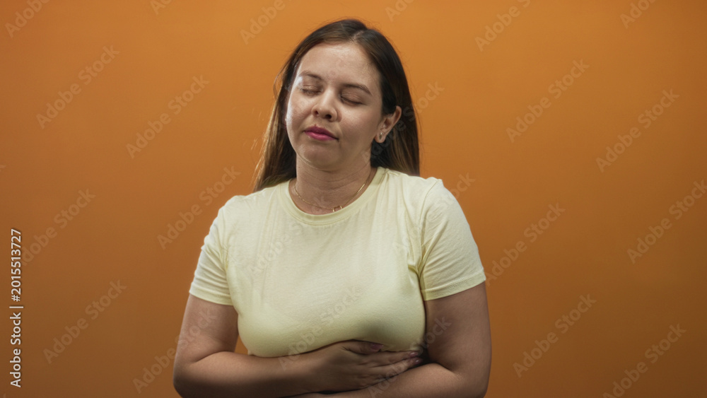 Obraz premium Young caucasian woman holding her stomach and touching her throat in an orange studio; discomfort nausea.
