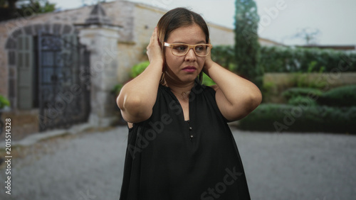 Photos Woman covering ears with hands at building entrance outdoors in front of stone gate and trimmed hedge, wearing glasses and sleeveless black top; noise annoyance