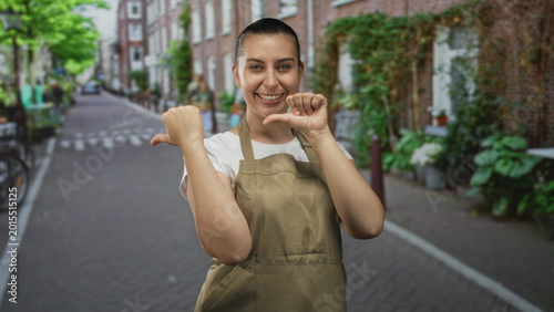 Foto Woman in olive apron pointing thumbs to chest while smiling on a narrow brick street with bicycles and potted plants; pride
