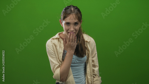 Konstfotografi Young woman covering mouth with hand and smiling slightly while facing camera in green studio; surprise youth innocence