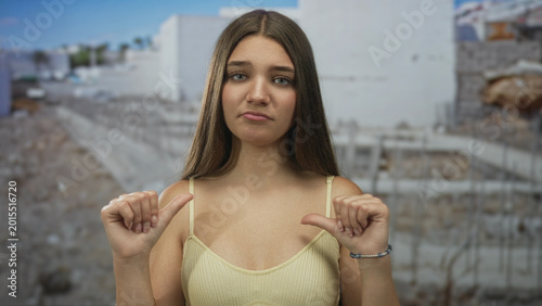 Photos Teenage woman stands amid concrete debris at sunlit building site pointing thumbs to chest for emphasis; confidence