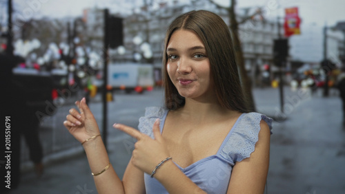 Photos Teenager caucasian girl in light blue top points finger to city street crossing while smiling at camera; playfulness