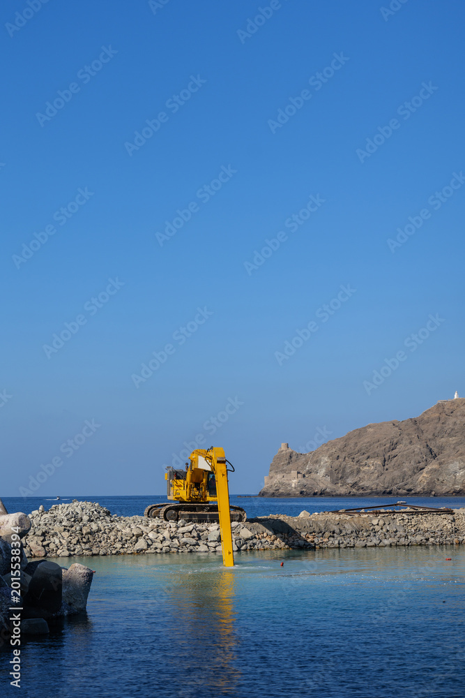 Fototapeta premium Coastal construction excavator working on rocky breakwater featuring marine construction, waterfront engineering and industrial coastal development