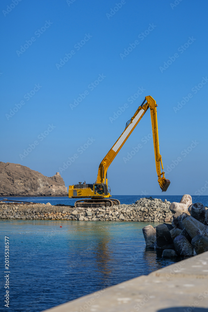 Fototapeta premium Marine infrastructure construction featuring excavator on stone breakwater, coastal engineering and waterfront industrial development