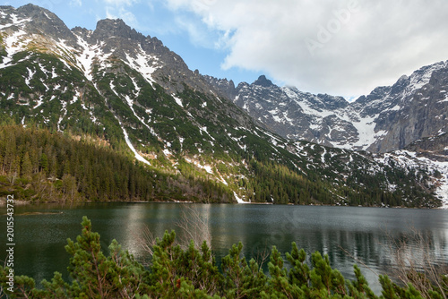 Scenic view of snow-capped mountains reflecting in a tranquil lake surrounded by lush green trees under a partly cloudy sky in a natural landscape