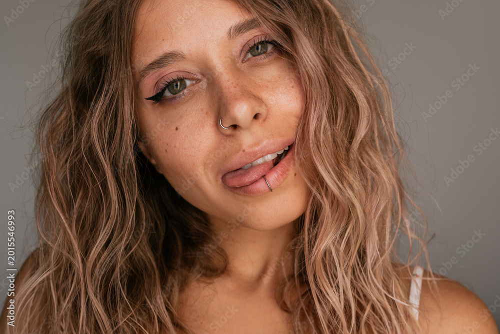 Naklejka premium Close-up portrait of young attractive blonde woman with wavy hair, freckles, winged eyeliner and fake lip piercing, playfully sticking out her tongue