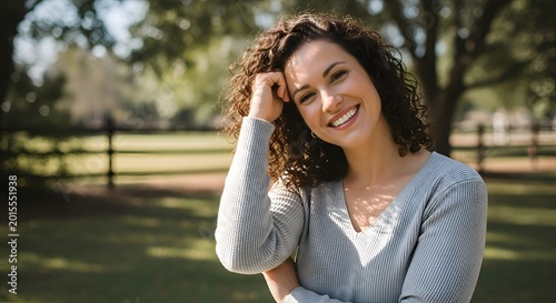 Radiant portrait of a smiling young woman with curly hair, enjoying a serene outdoor moment amidst lush greenery and a rustic wooden fence, embodying natural beauty.