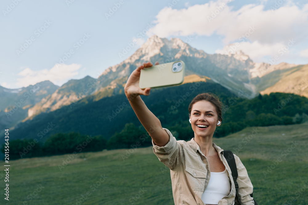 Fototapeta premium A cheerful woman takes a selfie in a scenic mountain meadow, casual jacket, sunny day. Lifestyle portrait capturing outdoor exploration and joyful travel moments, authentic, bright, modern.