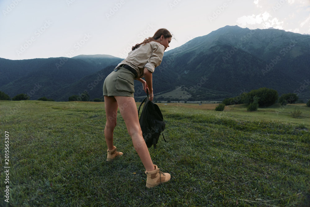 Fototapeta premium Young woman hiking across a lush meadow toward distant mountains, carrying a jacket and backpack, enjoying a breezy outdoor moment amid wide green scenery.