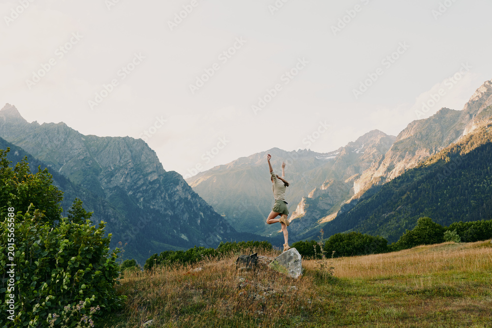 Naklejka premium Young woman leaps on a rock in a sunlit alpine meadow with distant rugged mountains, radiating freedom and joy in an outdoorsy, carefree moment