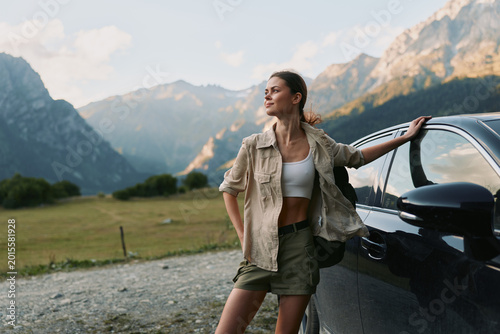 Fotografía A solo traveler, a young woman, leans on a dark car in a mountain landscape, wearing a white crop top, khaki shorts and a beige jacket, exuding casual adventure