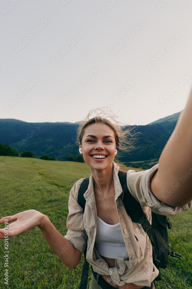 Fototapeta premium Smiling woman with backpack taking a selfie in an open meadow near mountains. Outdoor lifestyle, nature exploration, and travel mood. Bright natural light creates a warm vibe.