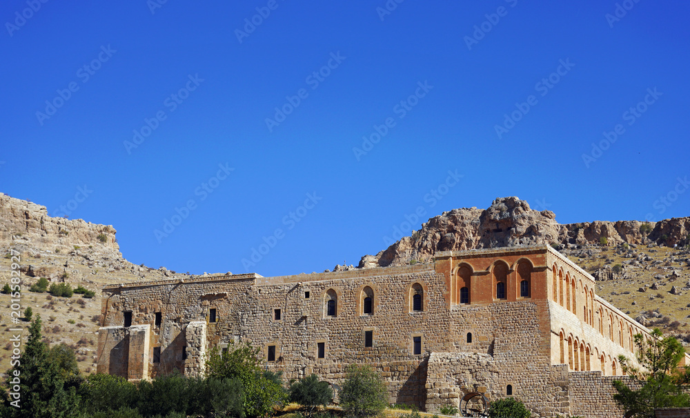 Obraz premium The historic stone facade of the ancient Deyrulzafaran Monastery (Mor Hananyo Monastery) stands at the base of arid, rugged mountains under a clear blue sky near Mardin, southeastern Turkey