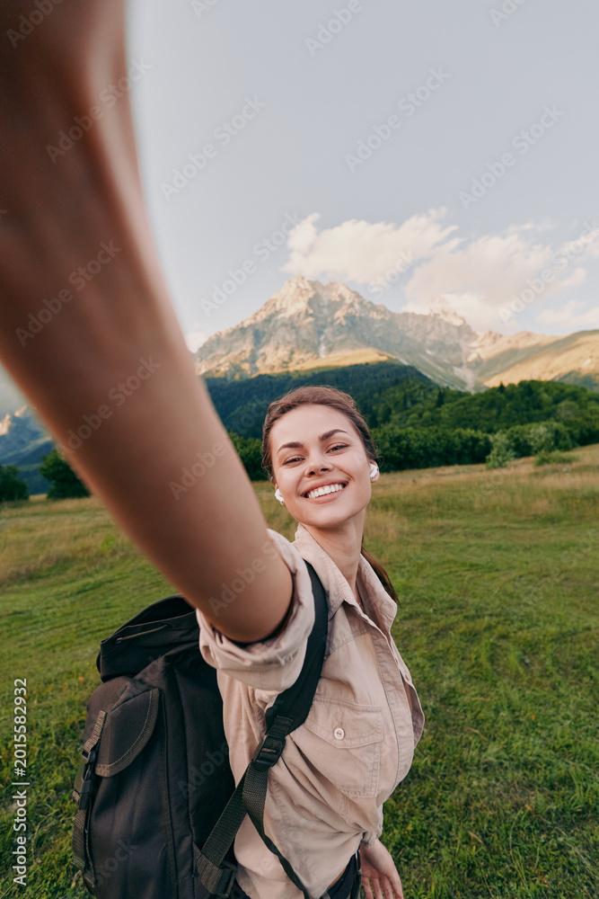 Naklejka premium Woman outdoors with backpack smiles for a selfie during a hike in a grassy valley beneath mountains, enjoying nature and travel in bright daylight, casual lifestyle
