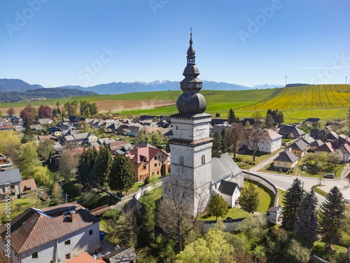 Aerial view of the historic St. Matthew's Church tower and traditional houses surrounded by green fields and distant mountains under a clear blue sky in Partizanska Lupca, Zilina Region, Slovakia.