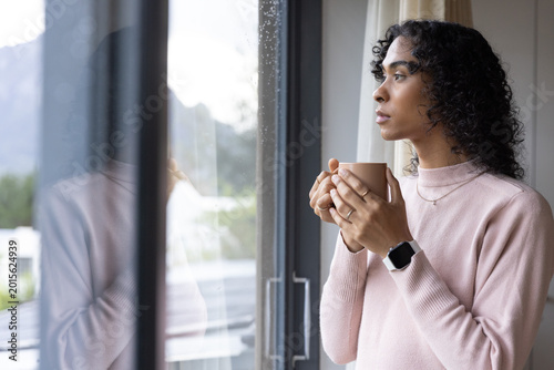 Non-binary person wearing pale pink pullover, gazing out window at home, holding mug, copy space