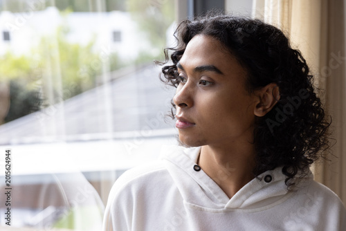 Non-binary person gazing toward right through large window at home, wearing white hoodie