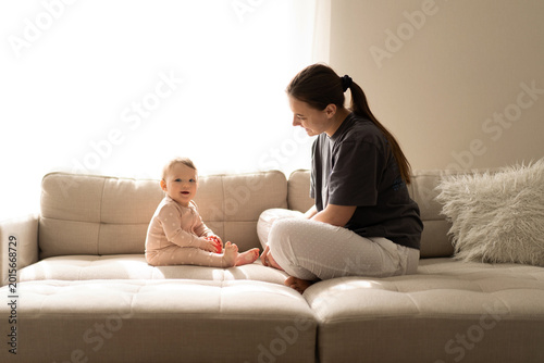 A mother sits on a sofa in a well-lit room and plays with her baby. Concept of teething period, baby care, early development, and healthy childhood life