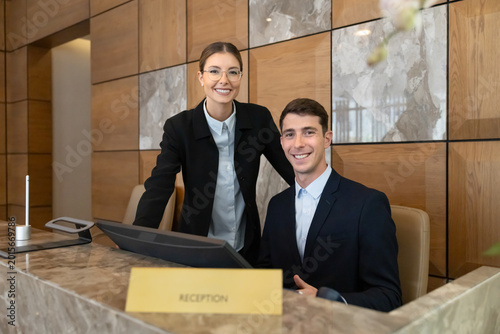 Portrait of professional hotel receptionists, colleagues posing behind front desk in modern hotel lobby, smiling, looking at camera, welcoming arriving guests. Hospitality services, tourism industry
