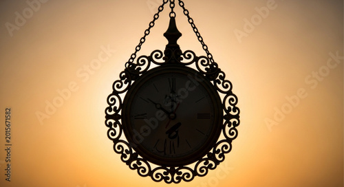 Antique ornate clock face hanging from chains against a sunset sky.