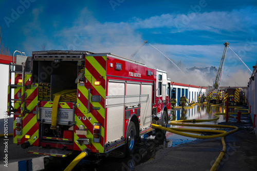 Obraz na płótnie Fire engine in the foreground with supply hose delivering water to aerial apparatus in the background spraying water on a burning storage facility as smoke rises into a blue sky
