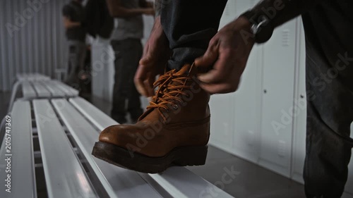 Close Up of Manual Laborer Resting Foot on Metal Bench, Carefully Tying Laces of Sturdy Protective Boots in Modern Warehouse or Factory Dressing Area. Concept of Industrial Gear and Readiness.