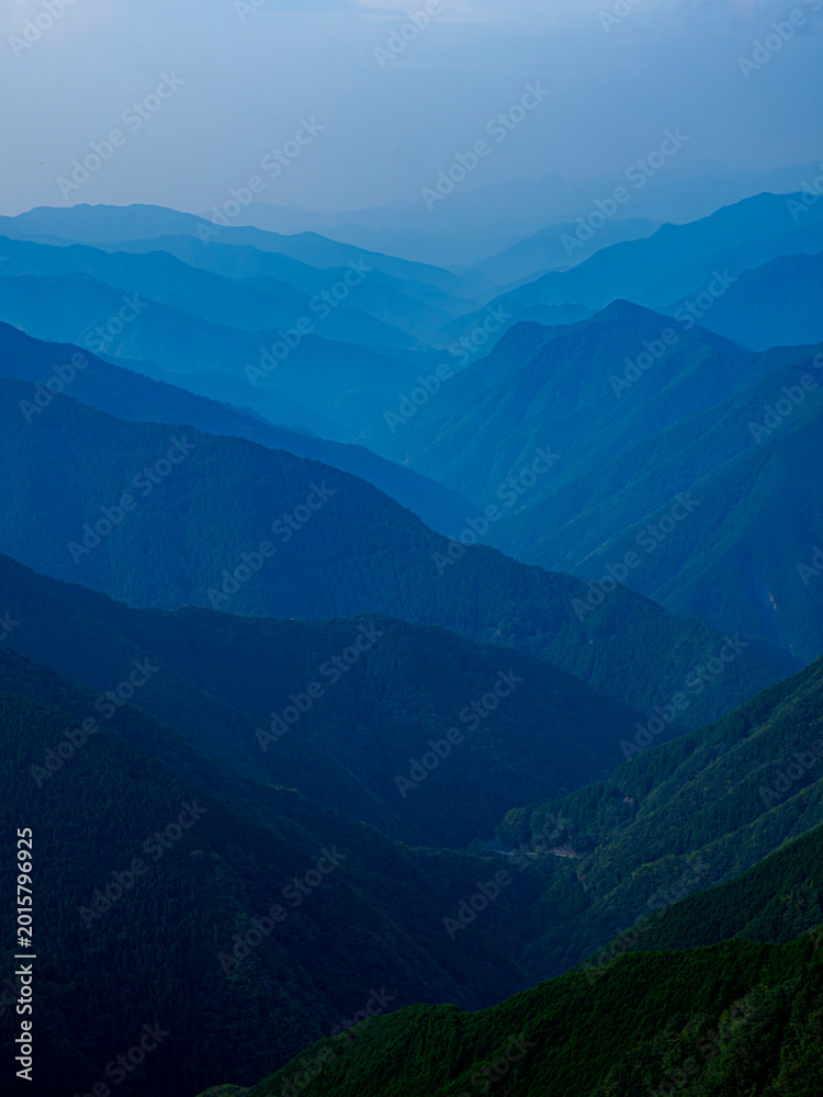 Fototapeta premium 夜明け。雄大な山岳地帯の風景。山の稜線。朝もや。ガス。霧。高山の森林がレイヤーのように折り重なる景色。自然風景。遠近感。空気遠近法のサンプル。縦構図