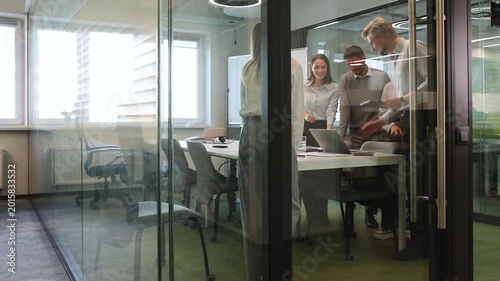 Group of business people standing together and discussing their work and projects, having a team meeting in an office, view from glass wall