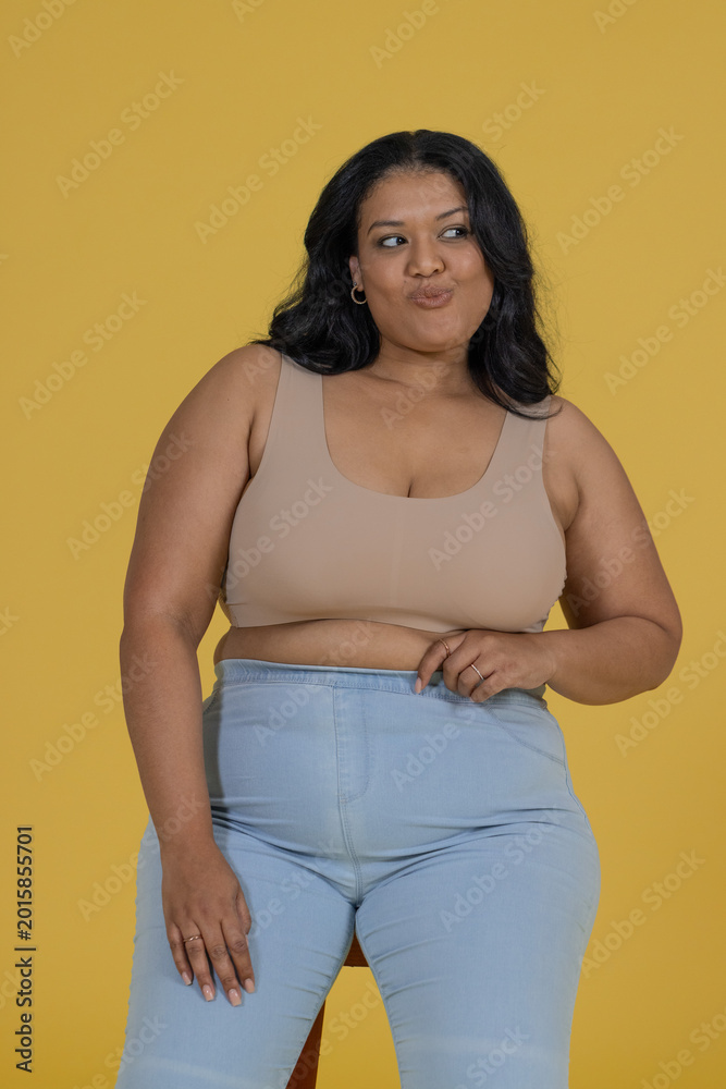 Obraz premium Woman posing on low stool in studio mustard backdrop croptop jeans hoops adjusting waist