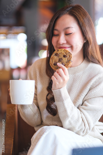 Vertical portrait image of a woman eating a piece of chocolate chip cookie and drinking coffee in cafe
