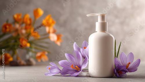 White bottle of lotion placed on table with purple flowers and orange flowers in background during bright daylight