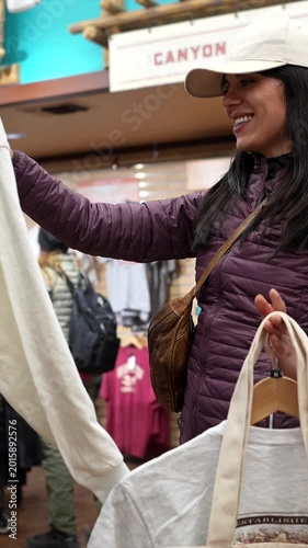 Smiling young woman wearing a baseball cap and puffy jacket browsing through clothes on hangers in a gift shop, looking for the perfect souvenir from her memorable travel experience