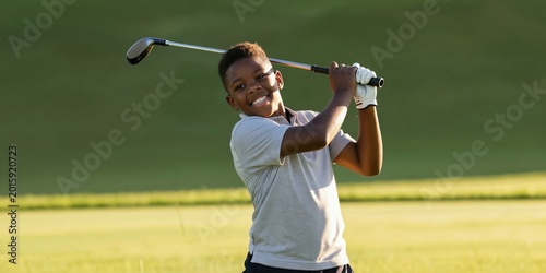 Young African american boy swings golf club on green course. Child enjoys outdoor sport game, smiles with happiness, focused on hitting ball. Healthy active pastime for kids.