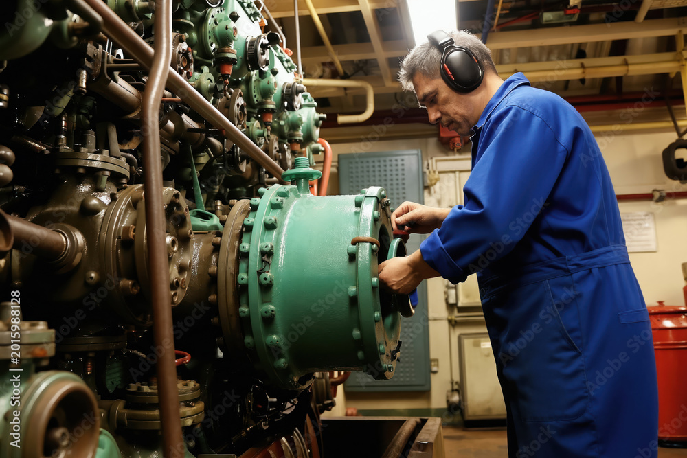 Obraz premium Marine engineer in blue overalls works on large ship engine. Man with headphones repairs complex machinery in engine room. Maritime industry worker maintains power plant.