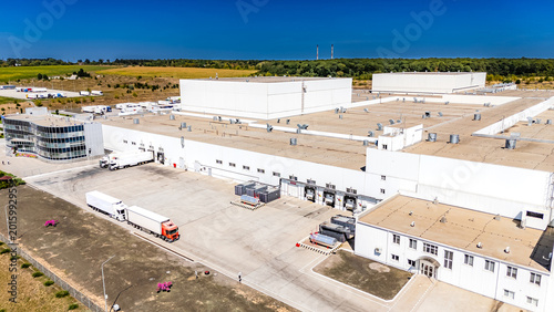 Logistics terminal with cargo trucks at loading bays. Perspective view of an industrial building exterior with semi trucks docked for loading and unloading operations.
