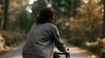 Person riding a bicycle on a road in a forest. the person is wearing a gray sweater and beige pants. the road is surrounded by trees with autumn foliage, and the ground is covered in fallen leaves.