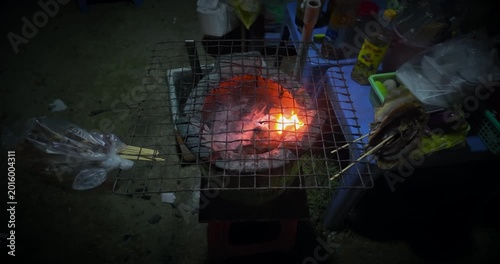 Slow motion close up of a traditional charcoal stove with a metal grill on top, featuring bright orange flames and smoke, set up for street food grilling at night