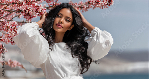 Beautiful woman posing under blooming cherry tree in spring. Stylish girl with long dark hair wearing white dress. Concept of natural female beauty, spring season and fashion portrait.