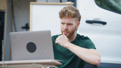 Thoughtful man working remotely on a laptop, sitting outside a camper van. Embracing digital nomad lifestyle and travel