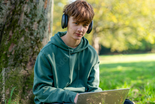 Foto Teen listens to music on headphones while using a laptop under a tree in a park