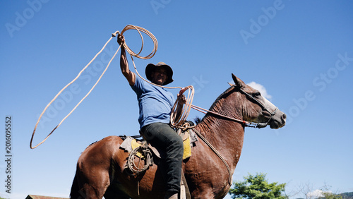 Foto Latino man wearing a hat and a blue shirt, riding a brown horse, holding a rope