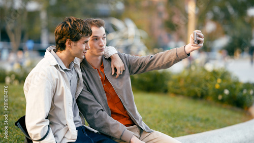 Принти на полотні Young gay men embracing while posing for a self portrait on a smartphone, enjoyi