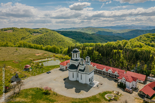 Aerial view of a serene monastery complex featuring a white church with dark domes, surrounded by rolling green mountains. 