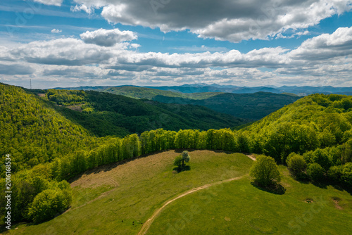 A scenic aerial view of rolling green hills and forested mountains under a partly cloudy sky. A winding dirt path leads through an open meadow, creating a peaceful and natural countryside atmosphere.