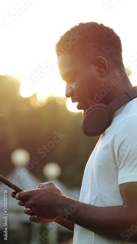 Black Man With Headphones Texting Smartphone During Sunset Urban Backlight. Portrayed As Student Checking Schedule, Musician Previewing Tracks, Commuter Streaming Podcast, Casual Friend Sharing