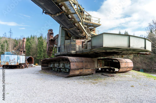 Large bucket wheel excavator with crawler tracks and long conveyor arm stands on a gravel yard near forest. The image shows open-pit mining machinery, industrial heritage, heavy equipment, steel const